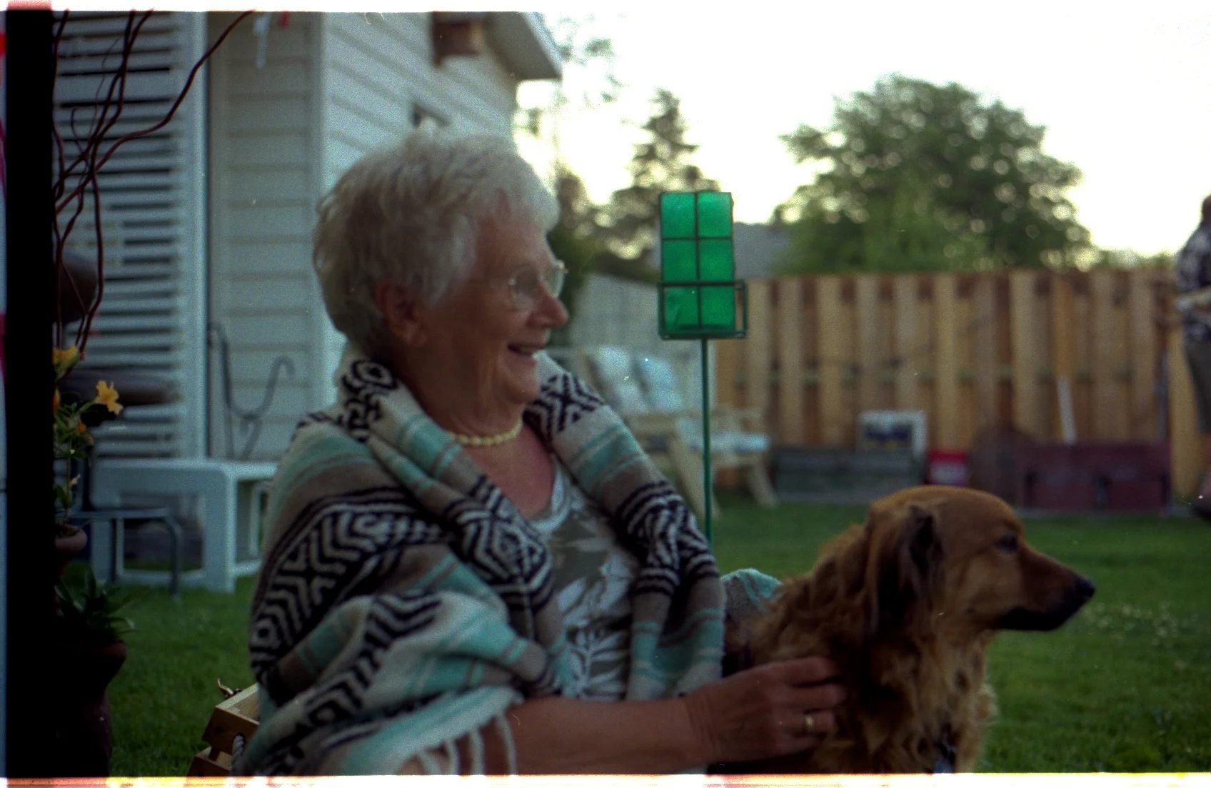 An elderly woman sitting outdoors in a garden, petting a golden retriever beside her.