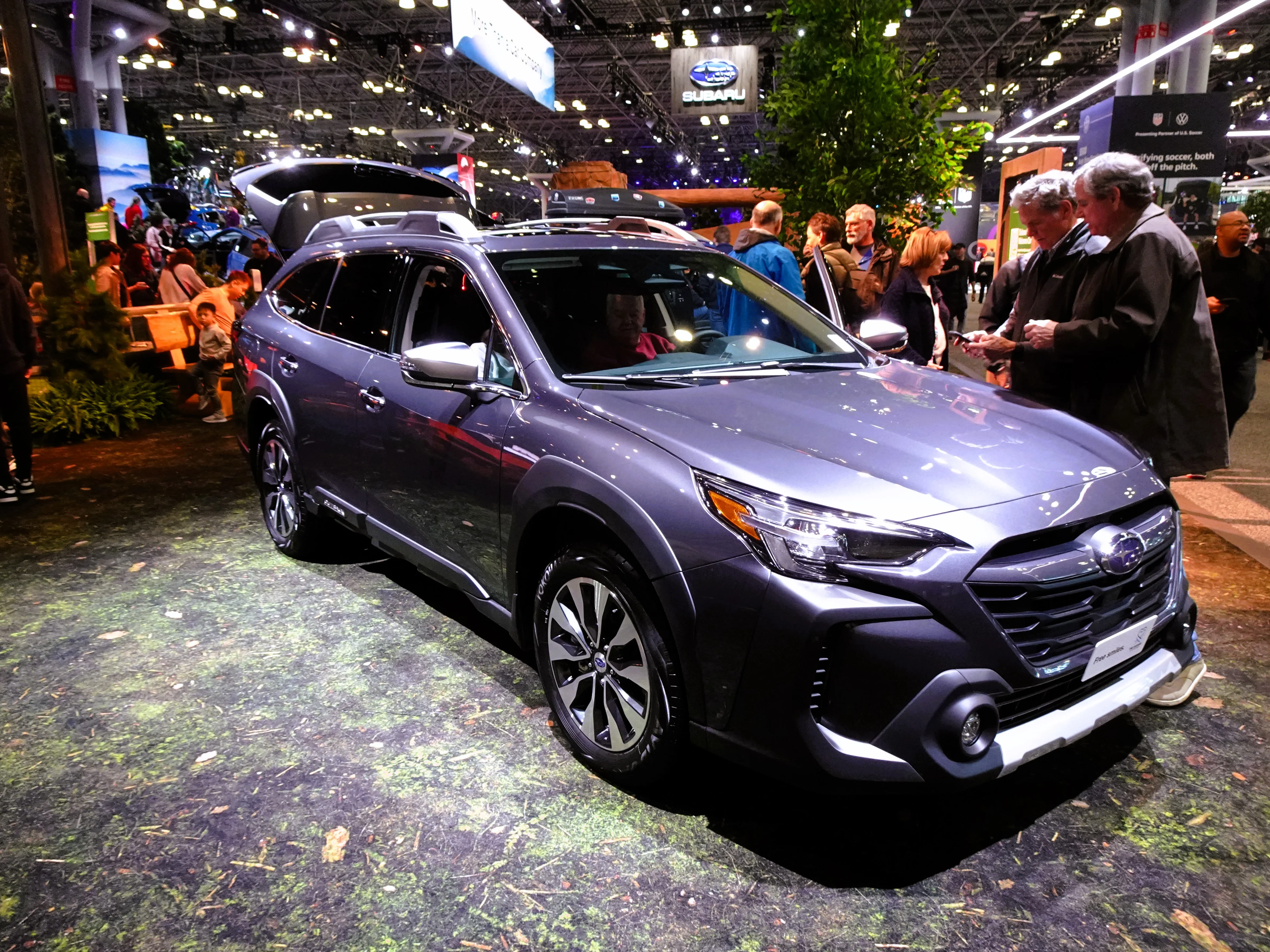 Grey Subaru Outback Touring XT wagon, current sixth generation, on an auto-show stand with a roof cargo box and its tailgate open.