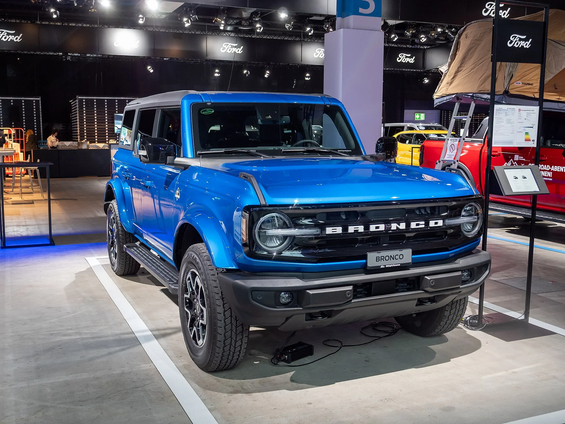 Blue four-door Ford Bronco Outer Banks on an auto-show stand with a rooftop tent displayed behind it, front three-quarter view.