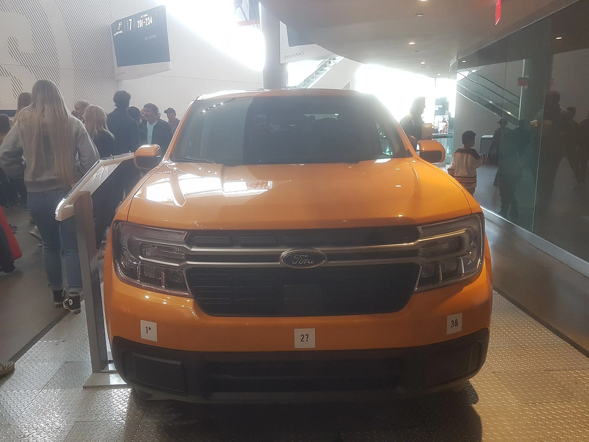 Orange Ford Maverick compact pickup, front view, showing its chrome Ford grille badge and slim split headlights on an indoor display stand