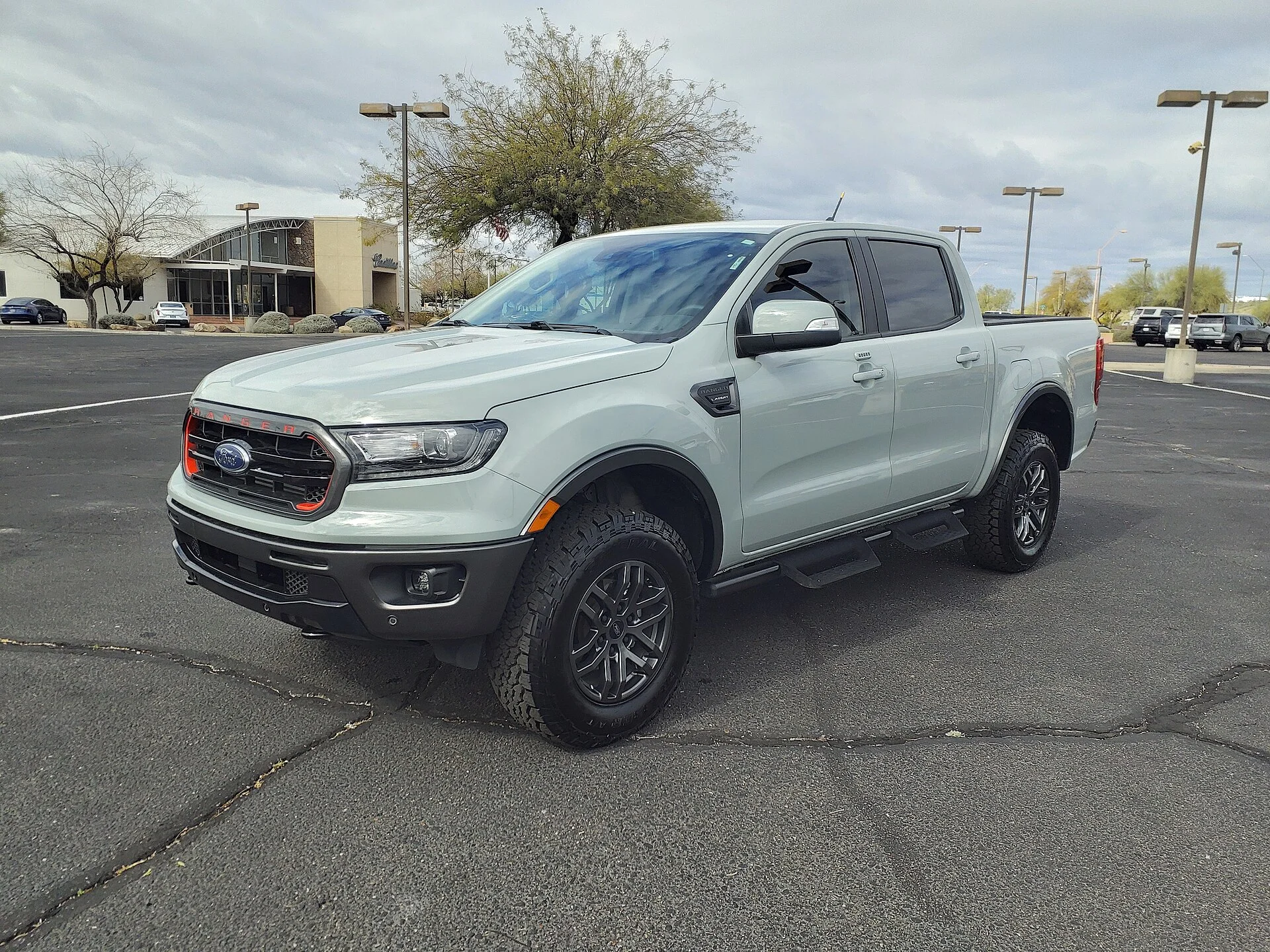Light sage-grey Ford Ranger Lariat crew-cab pickup in an empty parking lot, front three-quarter view showing its cargo bed, black grille with the Ford oval, and off-road tires