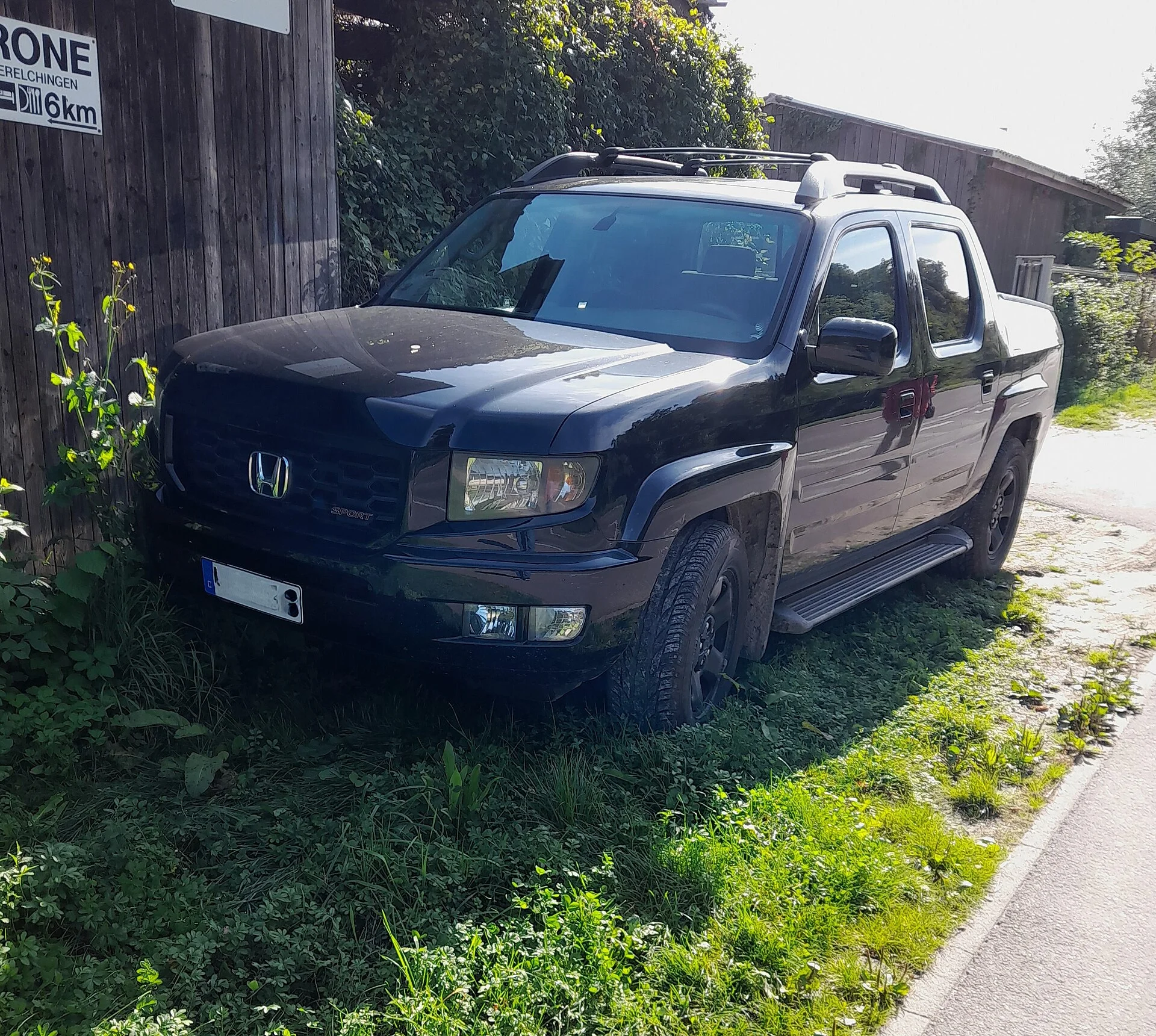 Black Honda Ridgeline Sport crew-cab pickup, front three-quarter view showing the Honda badge and short cargo bed, parked beside a wooden barn