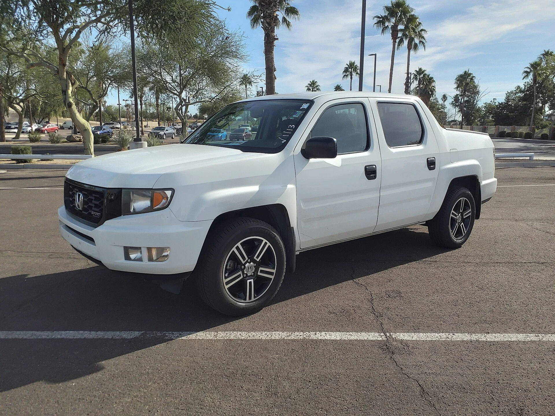White Honda Ridgeline crew-cab pickup in a parking lot with palm trees, front three-quarter view showing its cargo bed, black alloy wheels, and Honda grille