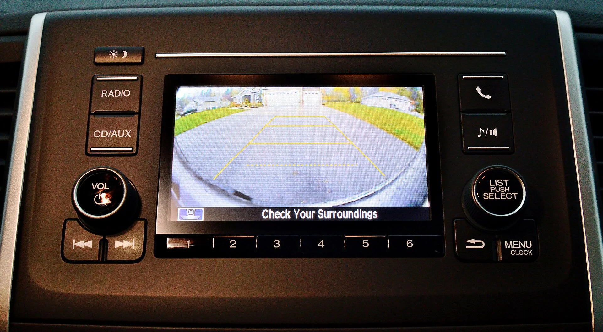 A car head unit screen showing the backup camera view with yellow distance guide lines over a driveway, labeled Check Your Surroundings