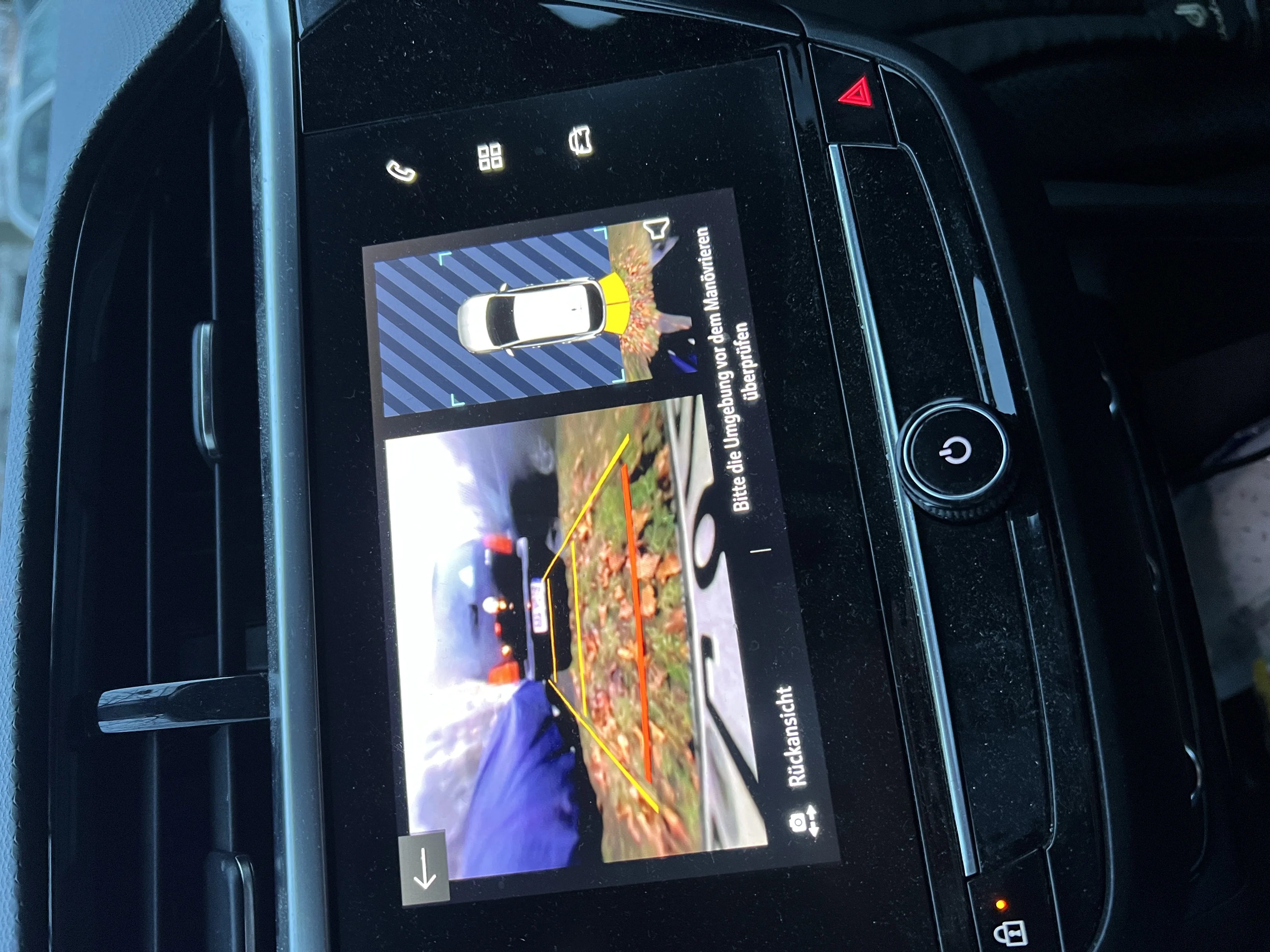 A car centre touchscreen showing the rear backup-camera view with coloured parking-guideline overlays and a top-down surround view during a reversing manoeuvre.