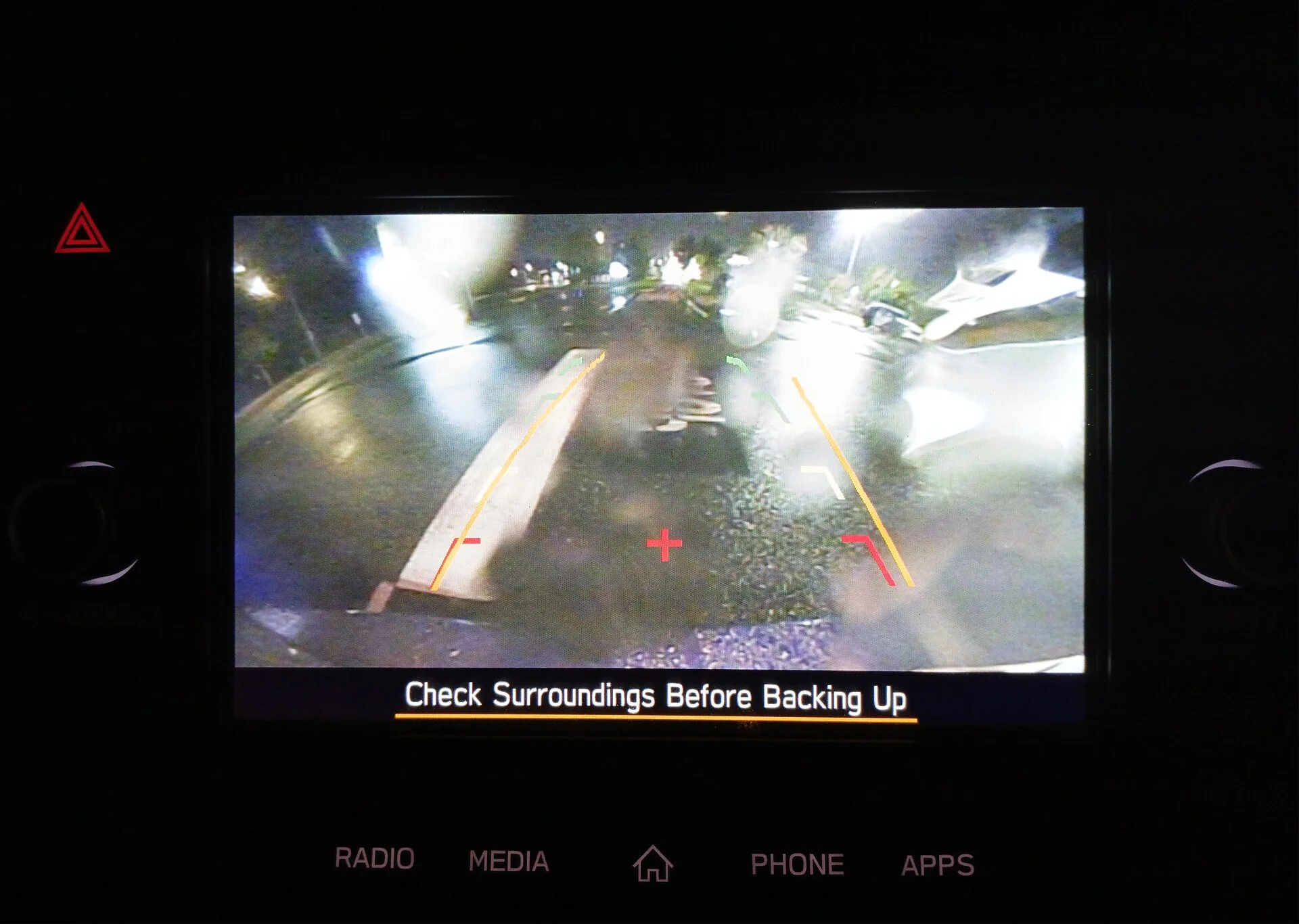 A car dashboard screen showing the rear backup-camera view at night with coloured parking-guideline overlays and a 'Check Surroundings Before Backing Up' prompt.
