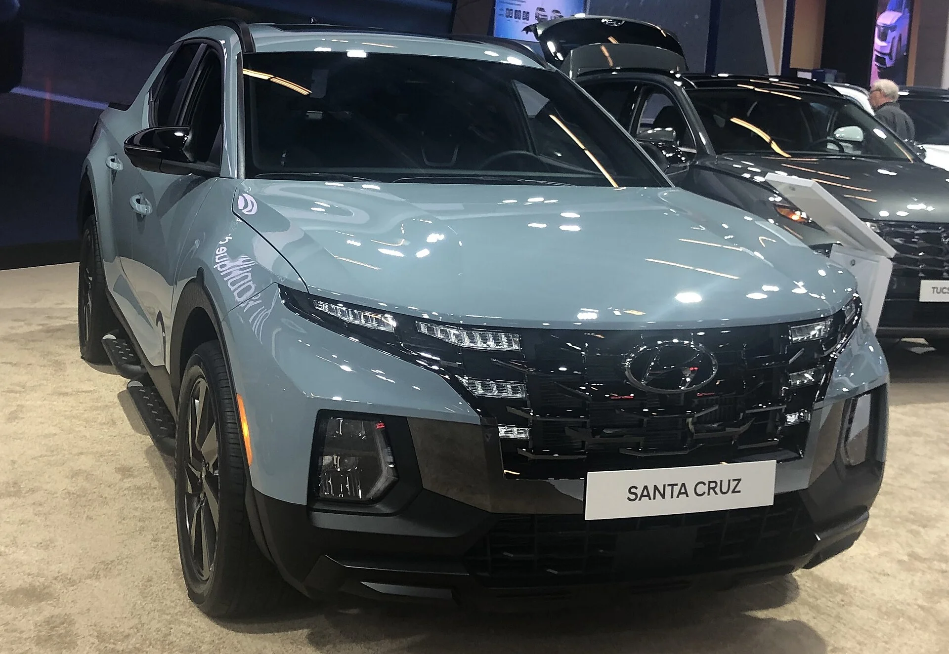 Grey-blue Hyundai Santa Cruz compact pickup, front three-quarter view showing the Hyundai badge and cascading grille, on an auto-show floor