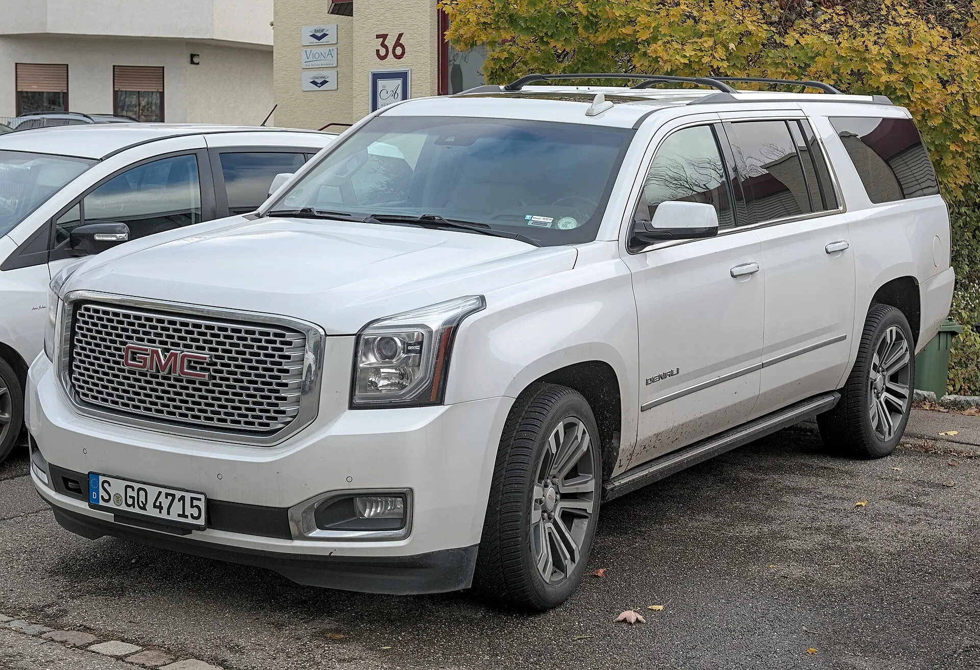 White GMC Yukon Denali full-size SUV parked on a street, front three-quarter view showing its large chrome grille, roof rails, alloy wheels, and Denali badging
