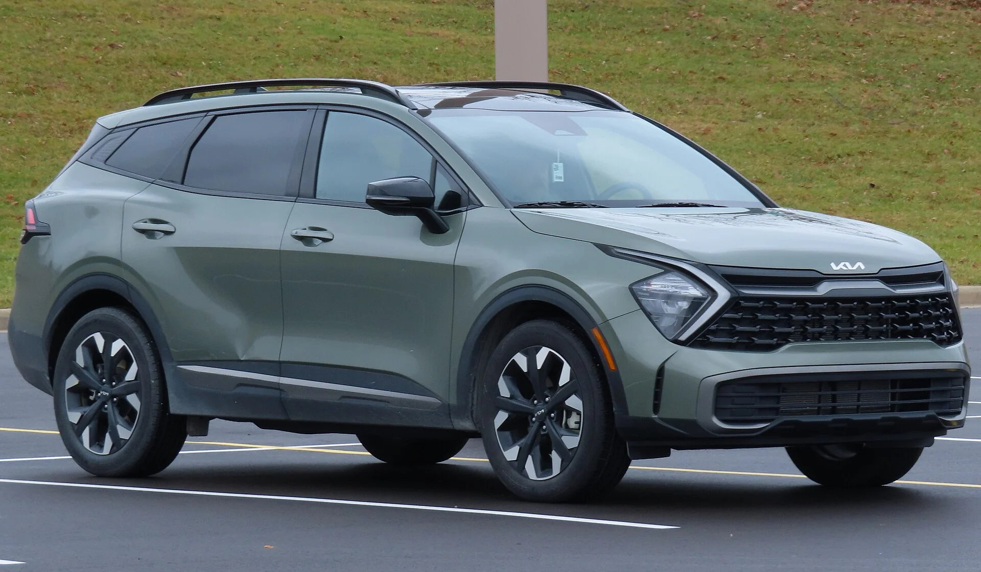 Sage-green Kia Sportage SUV in a parking lot, front three-quarter view showing its roof rails, black alloy wheels, and the Kia grille badge