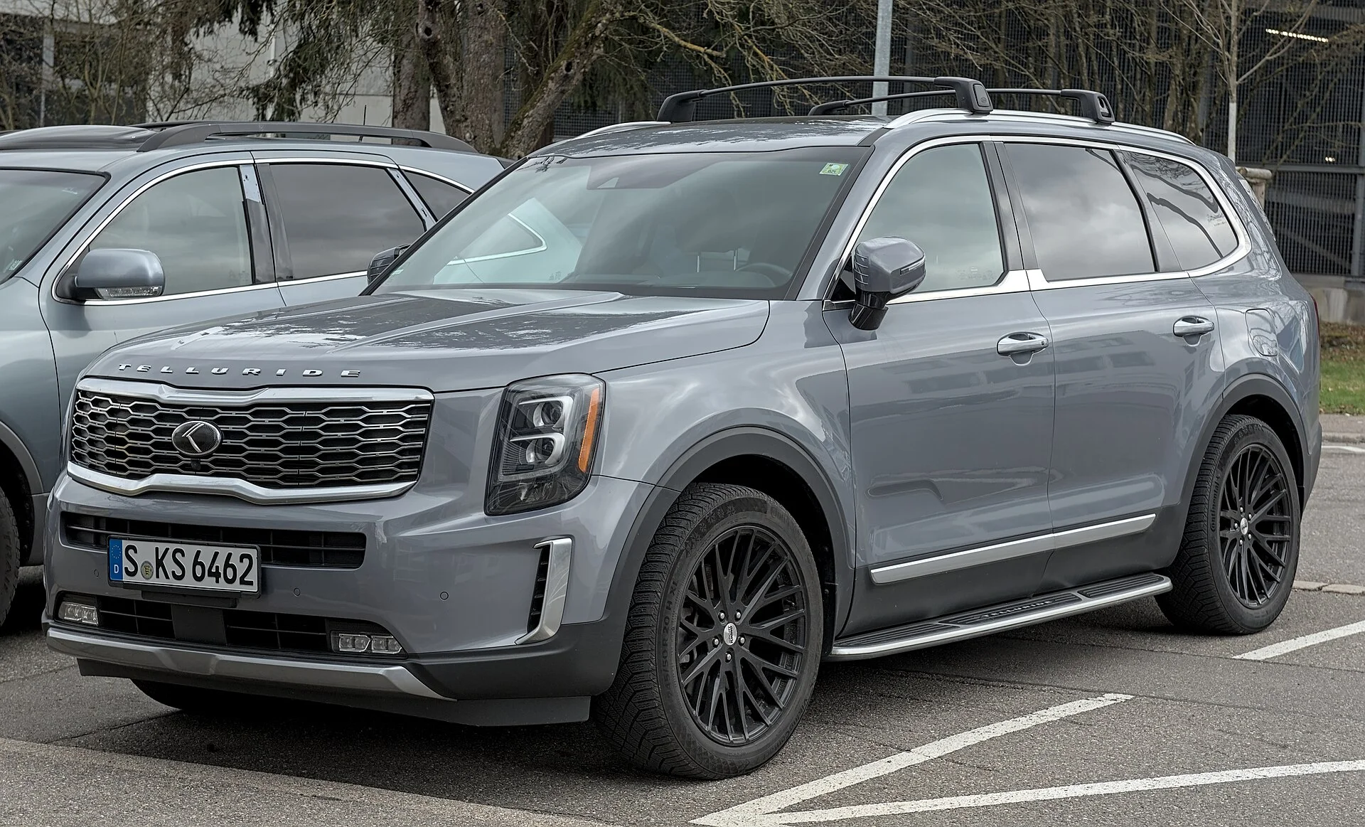 Grey Kia Telluride SUV in a parking lot, front three-quarter view showing its roof rails, running boards, black alloy wheels, and the Kia tiger-nose grille