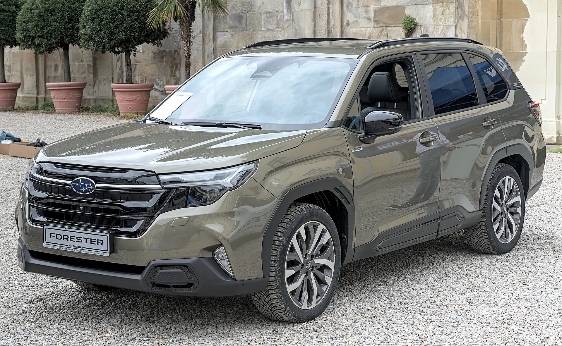 Olive-green Subaru Forester SUV parked on gravel in front of a stone wall, front three-quarter view showing its roof rails, alloy wheels, and Subaru grille badge
