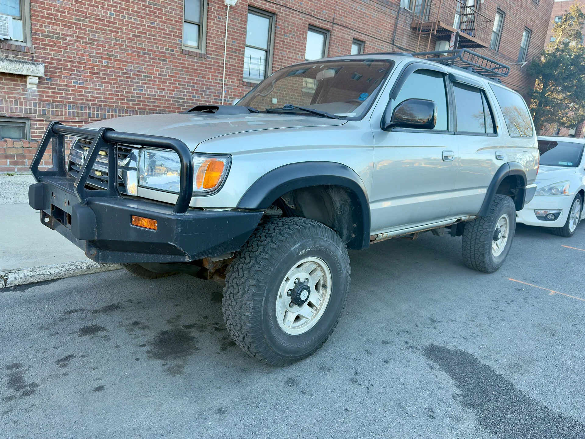 Silver older-generation Toyota 4Runner SUV fitted with a black brush guard and off-road tires, parked on a street beside a brick building, front three-quarter view