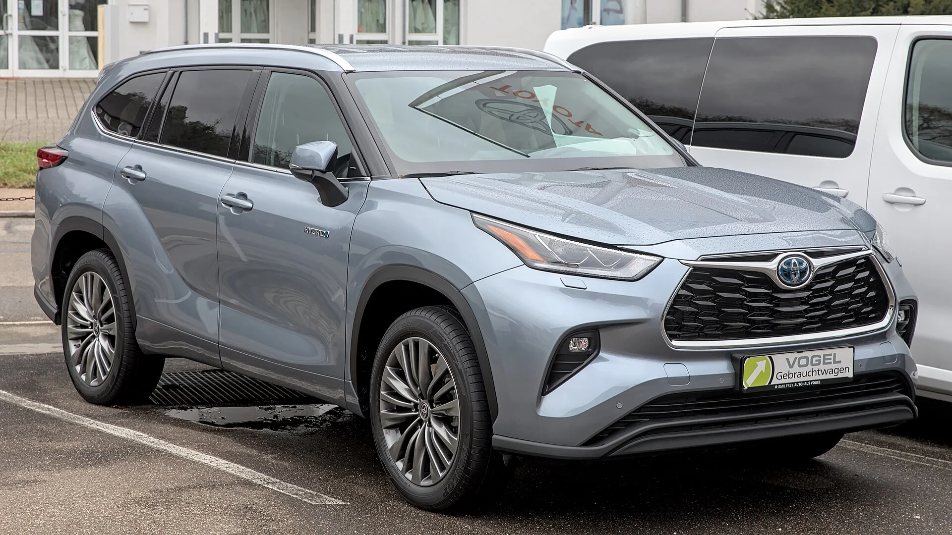 Silver-blue Toyota Highlander Hybrid SUV at a dealer lot, front three-quarter view showing its roof rails, alloy wheels, hybrid badge, and Toyota grille
