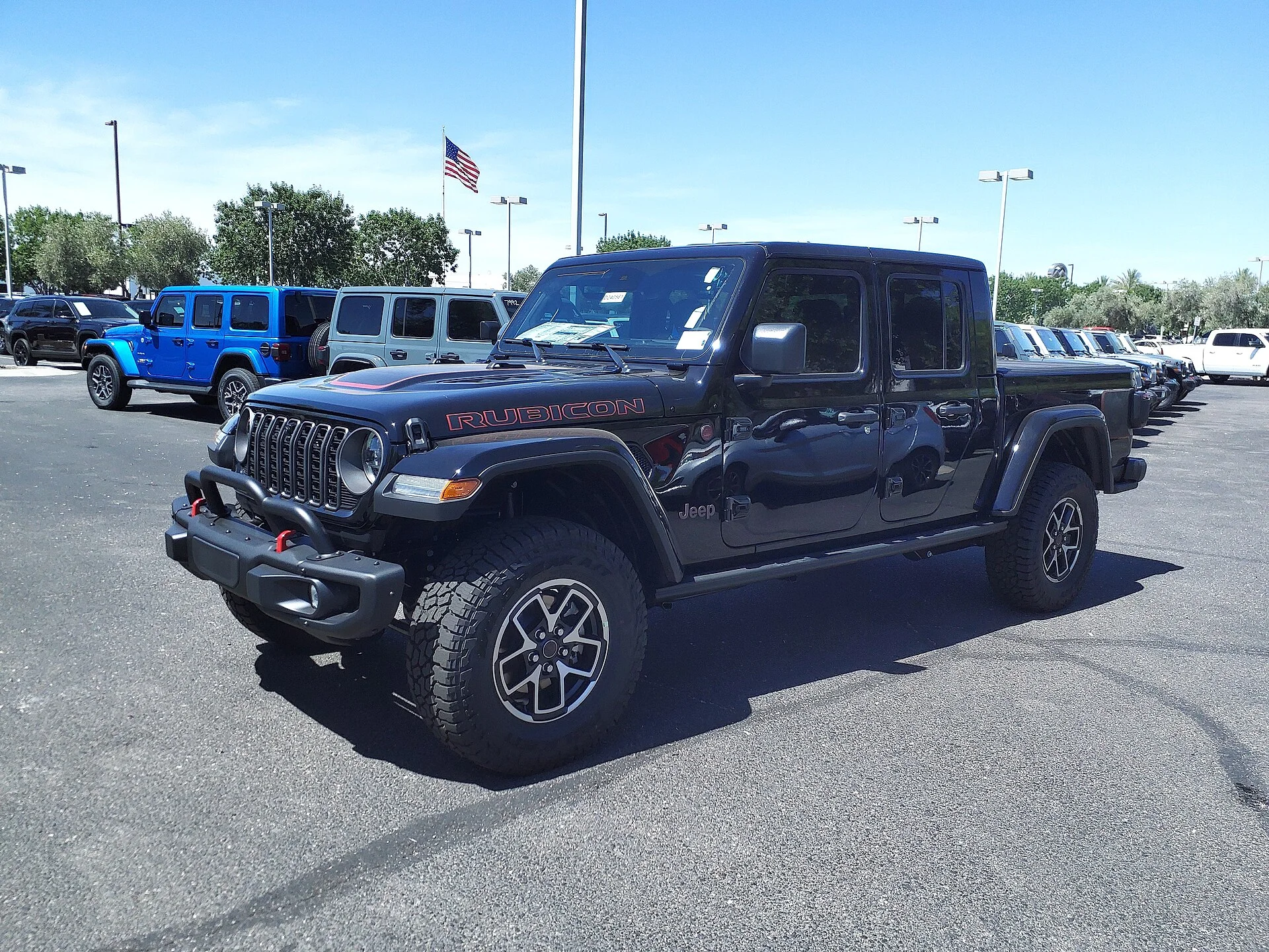 Dark grey Jeep Gladiator Rubicon pickup on a sunny dealer lot, front three-quarter view showing its seven-slot grille, RUBICON hood decal, short cargo bed, and off-road tires