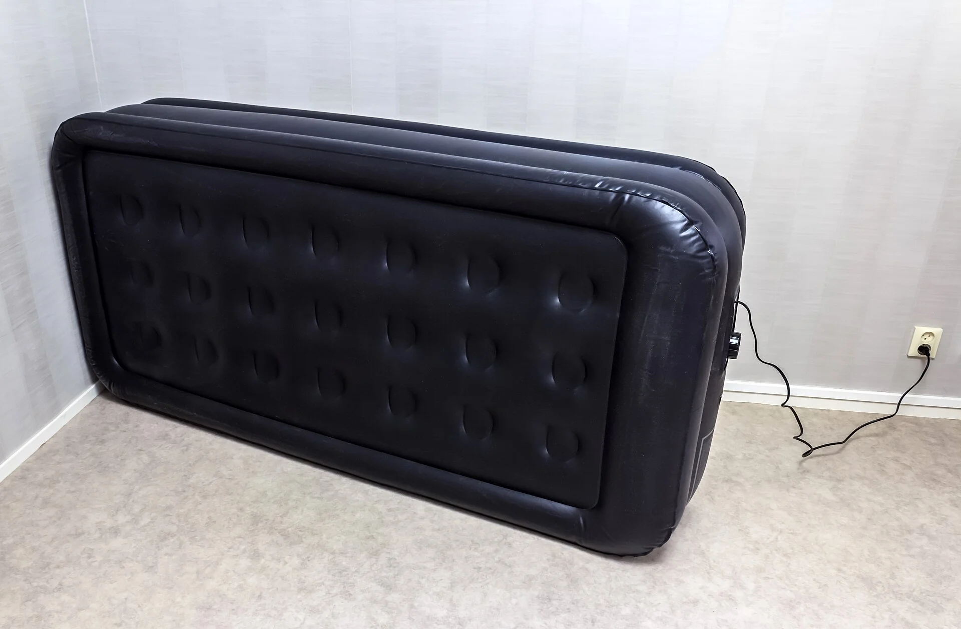 A black inflatable air mattress standing on its side, partly deflated, beside a wall power socket.