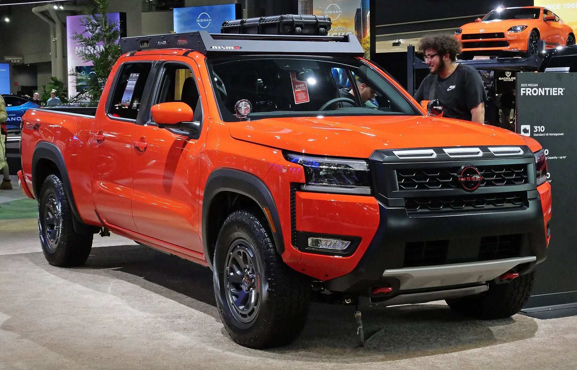 Orange Nissan Frontier Pro-4X crew-cab pickup fitted with a black roof rack, on display at an auto show, front three-quarter view showing its cargo bed, black grille, and off-road tires