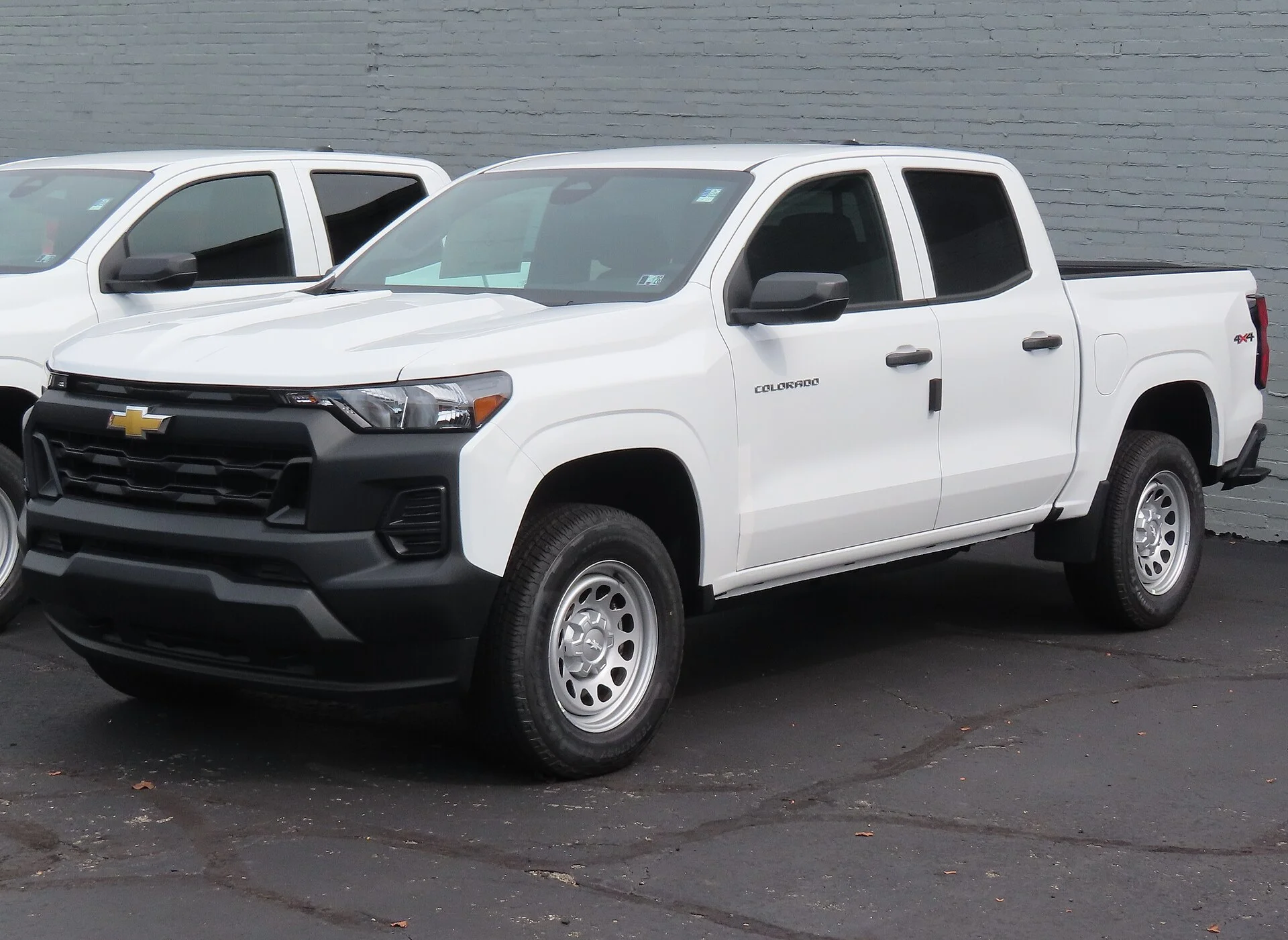 White Chevrolet Colorado WT crew-cab pickup beside a grey brick wall, front three-quarter view showing its cargo bed, black grille, steel wheels, and Colorado badging