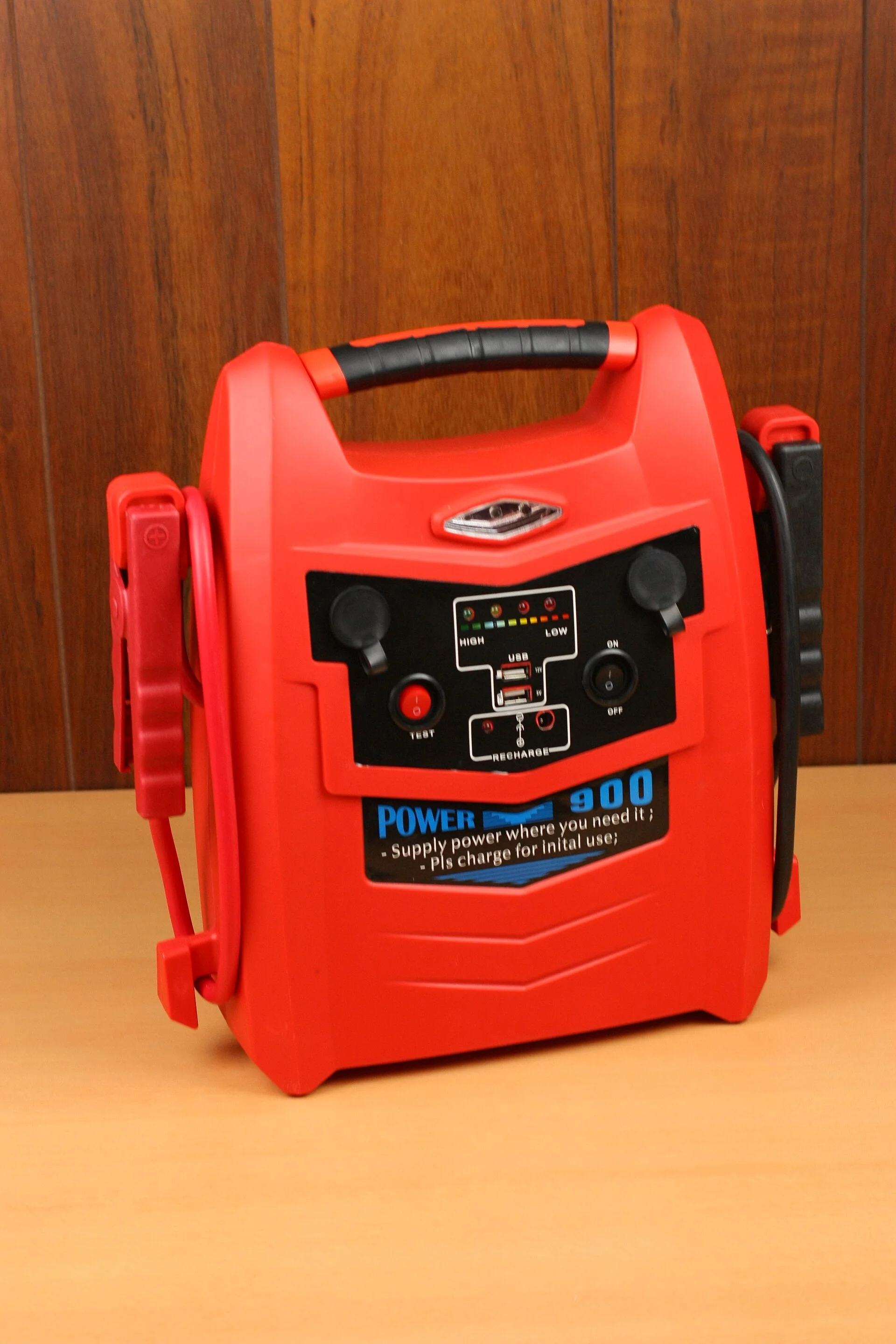A red portable jump-starter battery pack with attached jumper-cable clamps, a charge-level gauge and USB ports, on a desk.