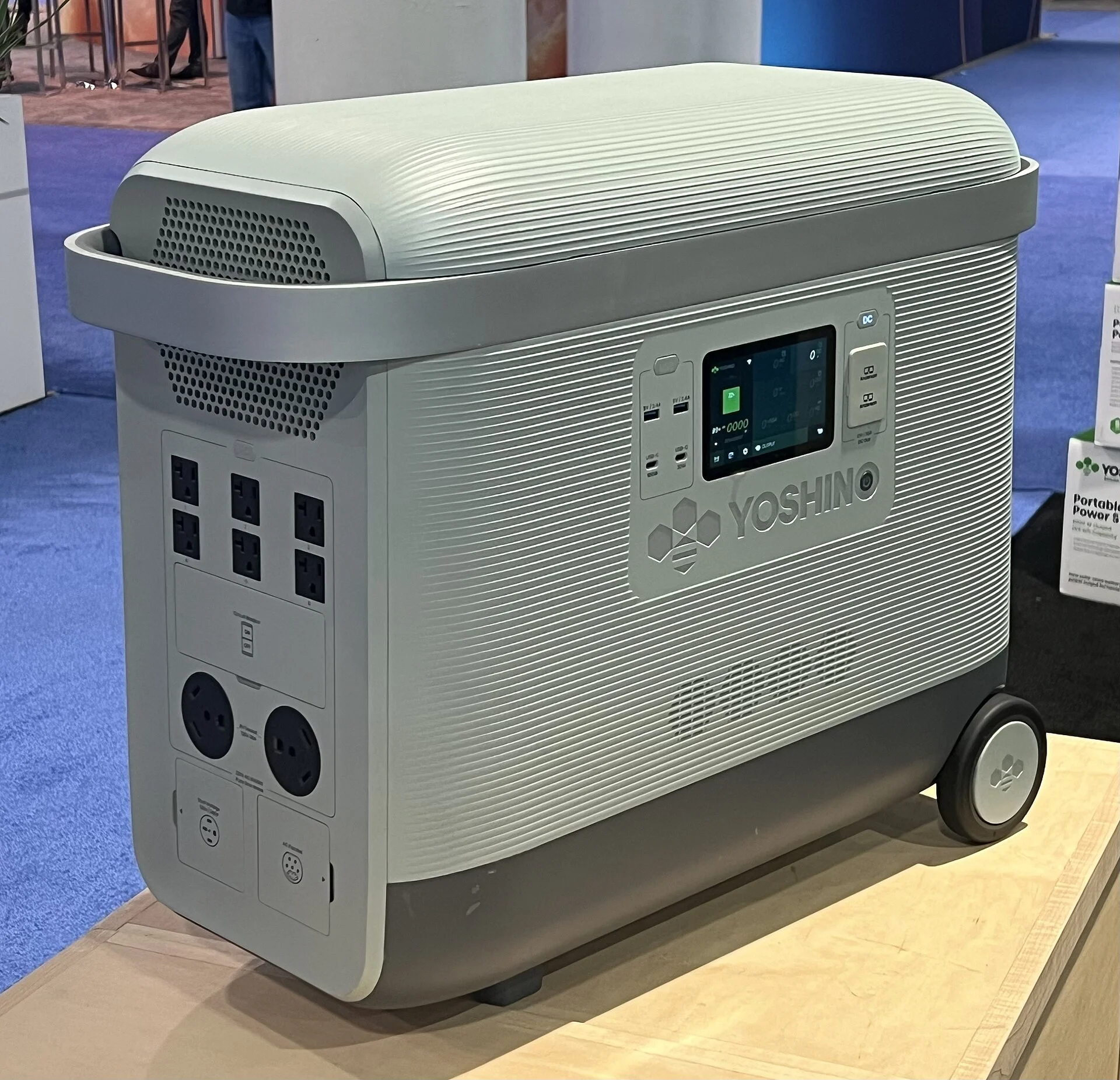 Light grey Yoshino B6000 portable power station on wheels, three-quarter view showing its side ventilation grilles, AC outlets, and display