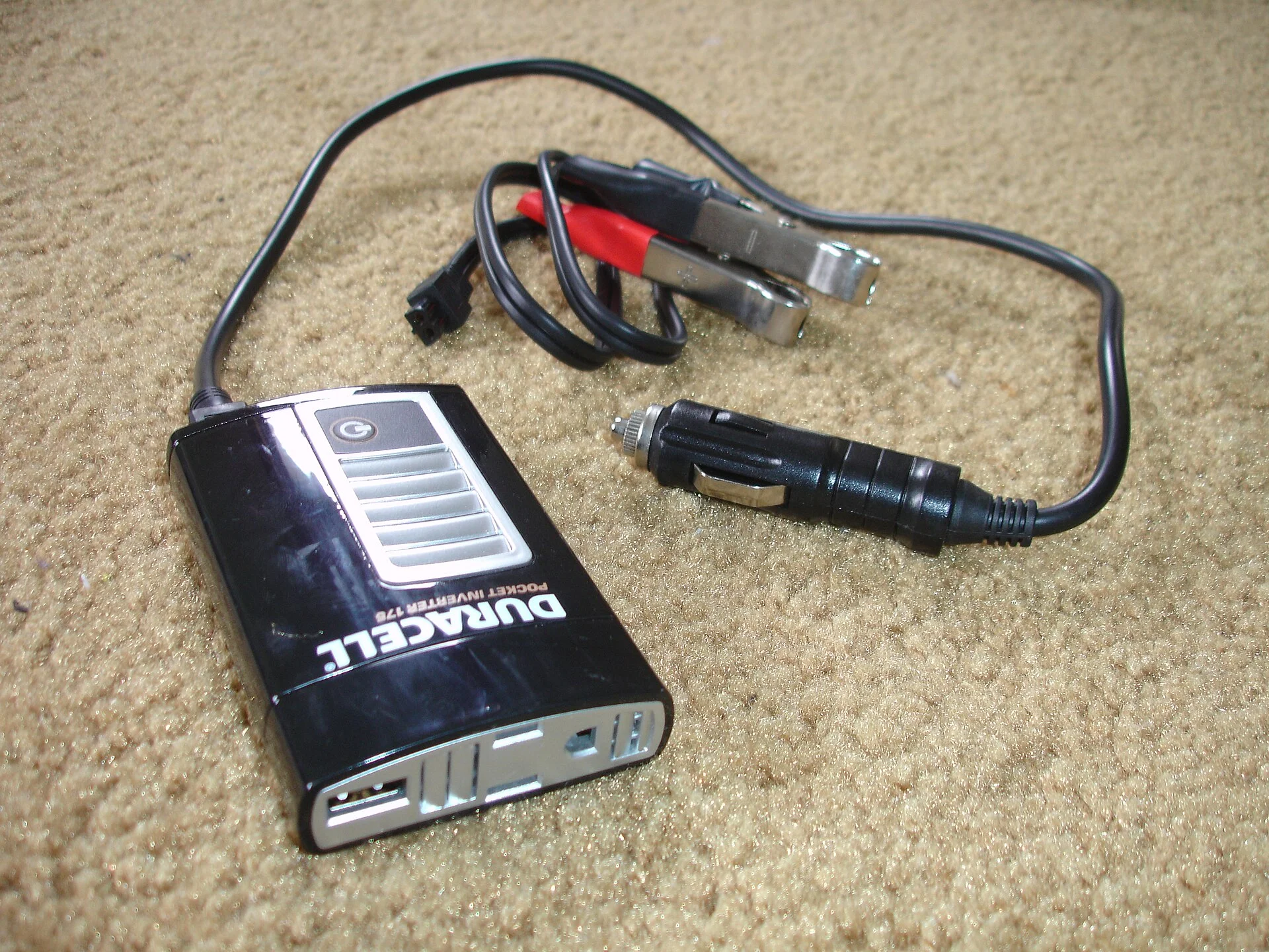 Small black pocket power inverter with a 12-volt cigarette-lighter plug and red-and-black battery clip cable, resting on sand