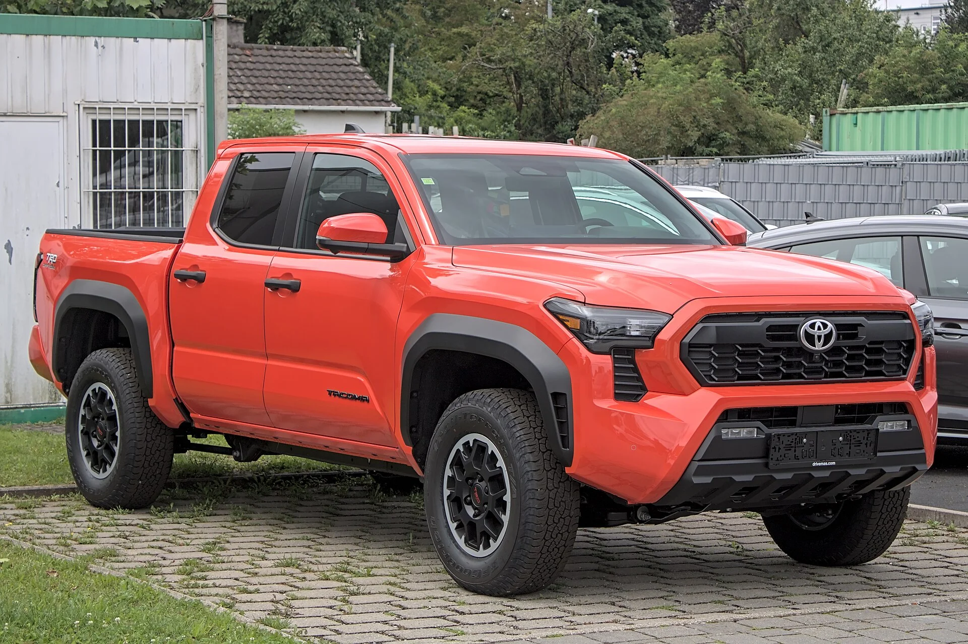 Bright orange 2024 Toyota Tacoma TRD Off Road double-cab pickup parked on grass, front three-quarter view showing its cargo bed, black fender flares, off-road tires, and Tacoma badging