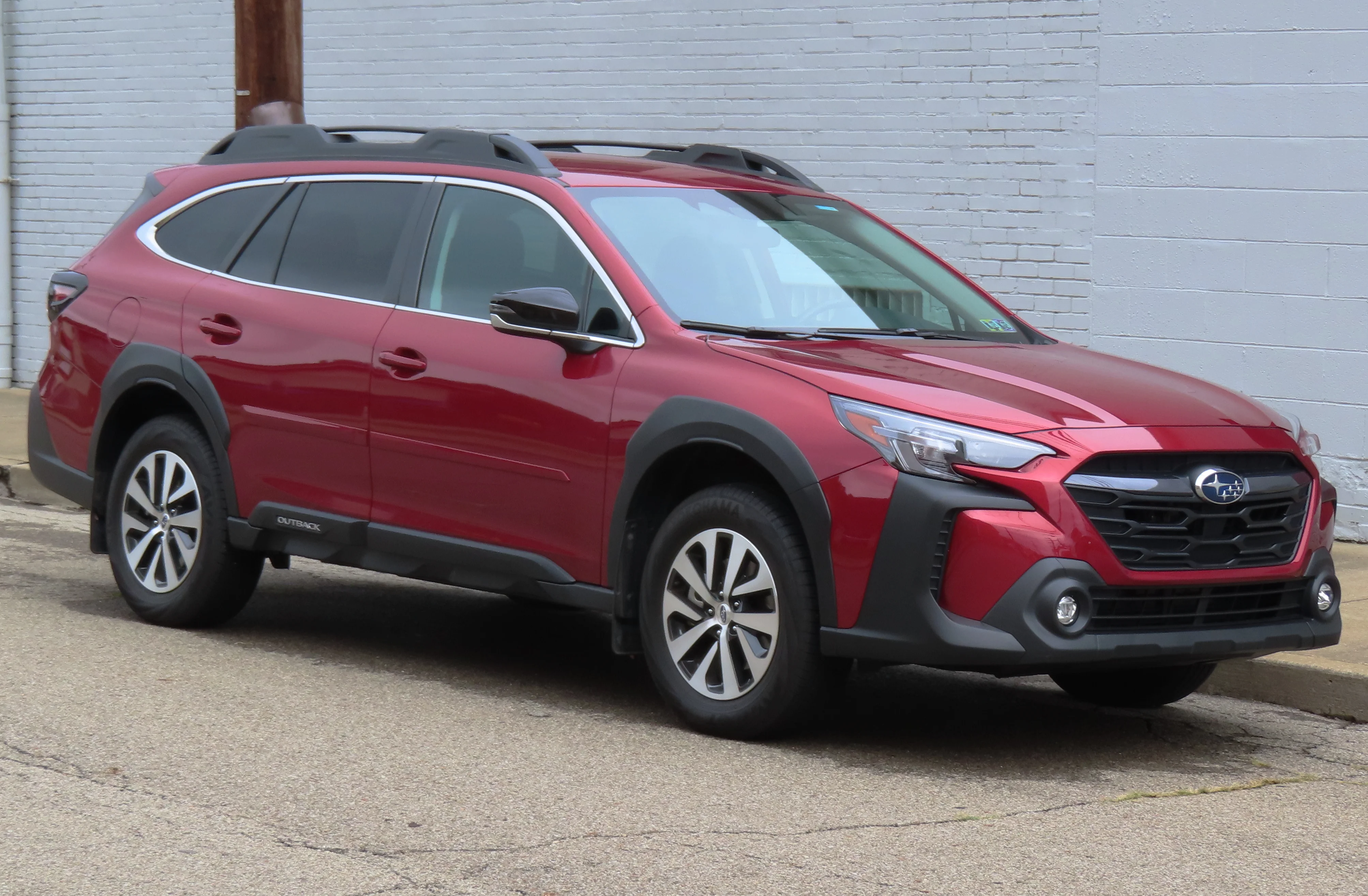 Red Subaru Outback Premium wagon, current sixth-generation, with black roof rails, front three-quarter view beside a brick wall.