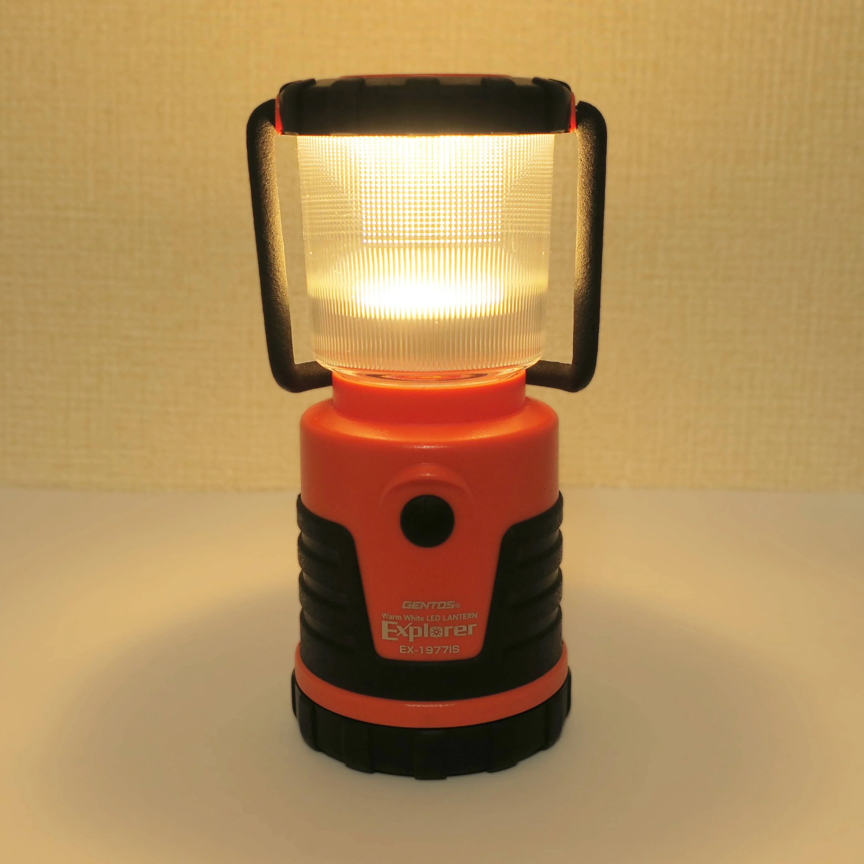 An orange-and-black LED camping lantern switched on and glowing, showing the warm light output judged for campsite use.