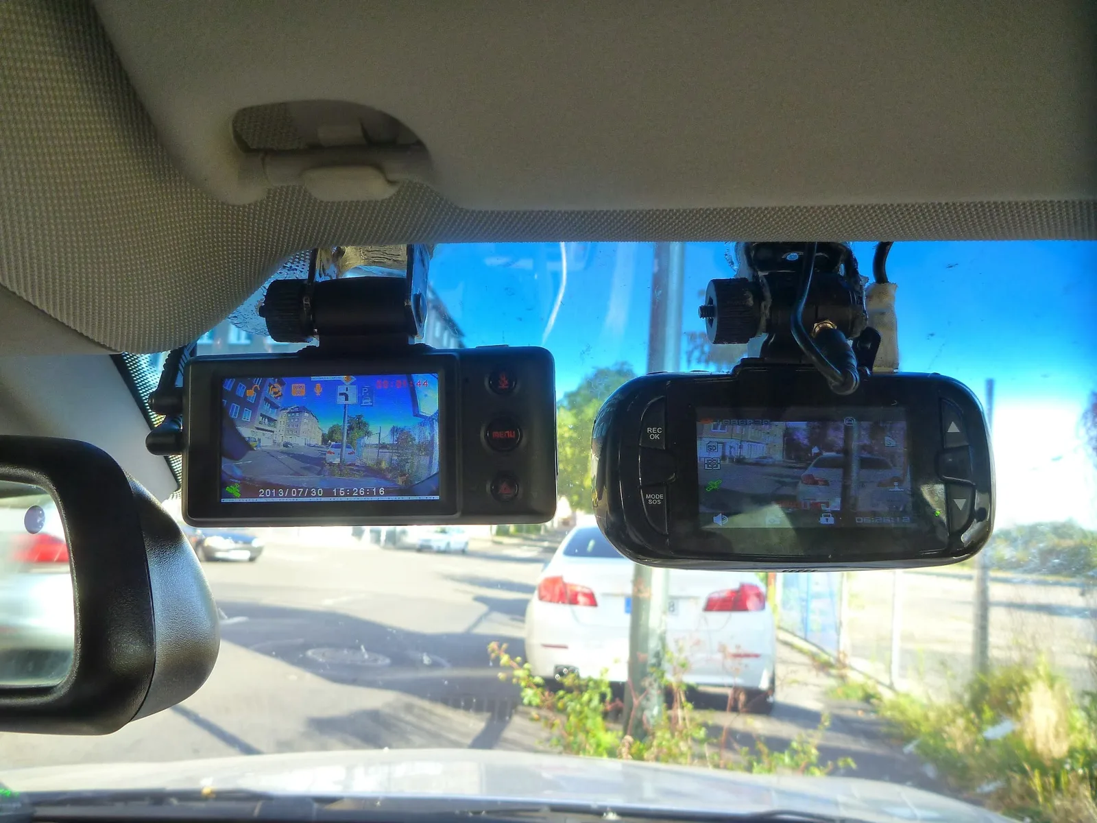 Two dash cams mounted side by side on a car windshield, both screens showing live footage, with a MENU button and a 2013/07/30 15:26:16 timestamp visible, relevant to adjusting a dash cam's date and time settings.