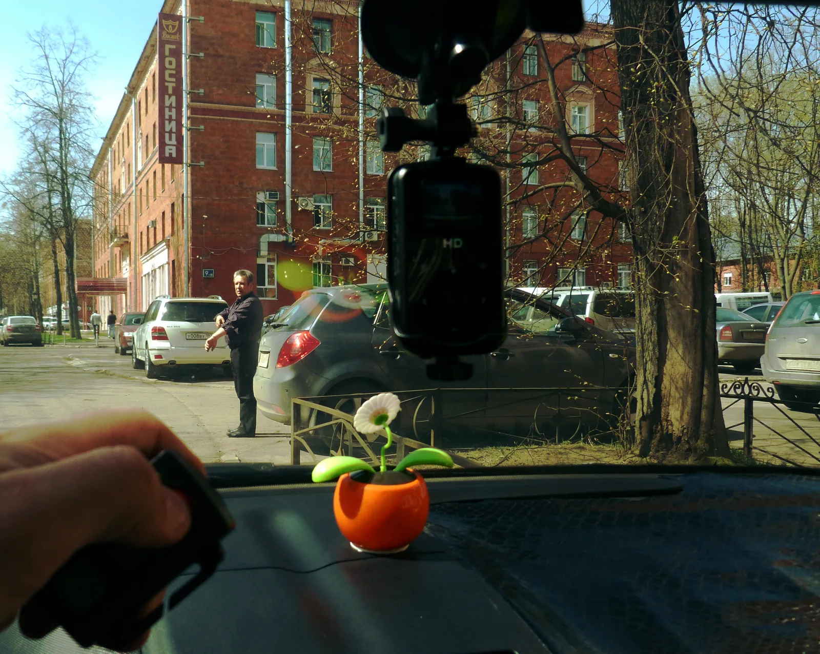 View from inside a parked car looking through the windshield at a street, with a black dashcam mounted on the glass in the foreground, showing a typical windshield dashcam installation.
