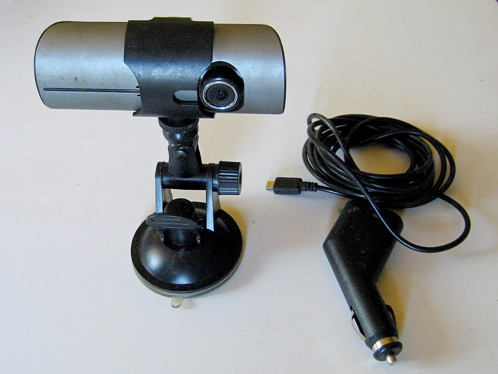 A dual-lens dashcam on a suction-cup windshield mount next to its coiled 12-volt car charger cable, shown on a plain background, illustrating a dashcam unit and how it is powered.