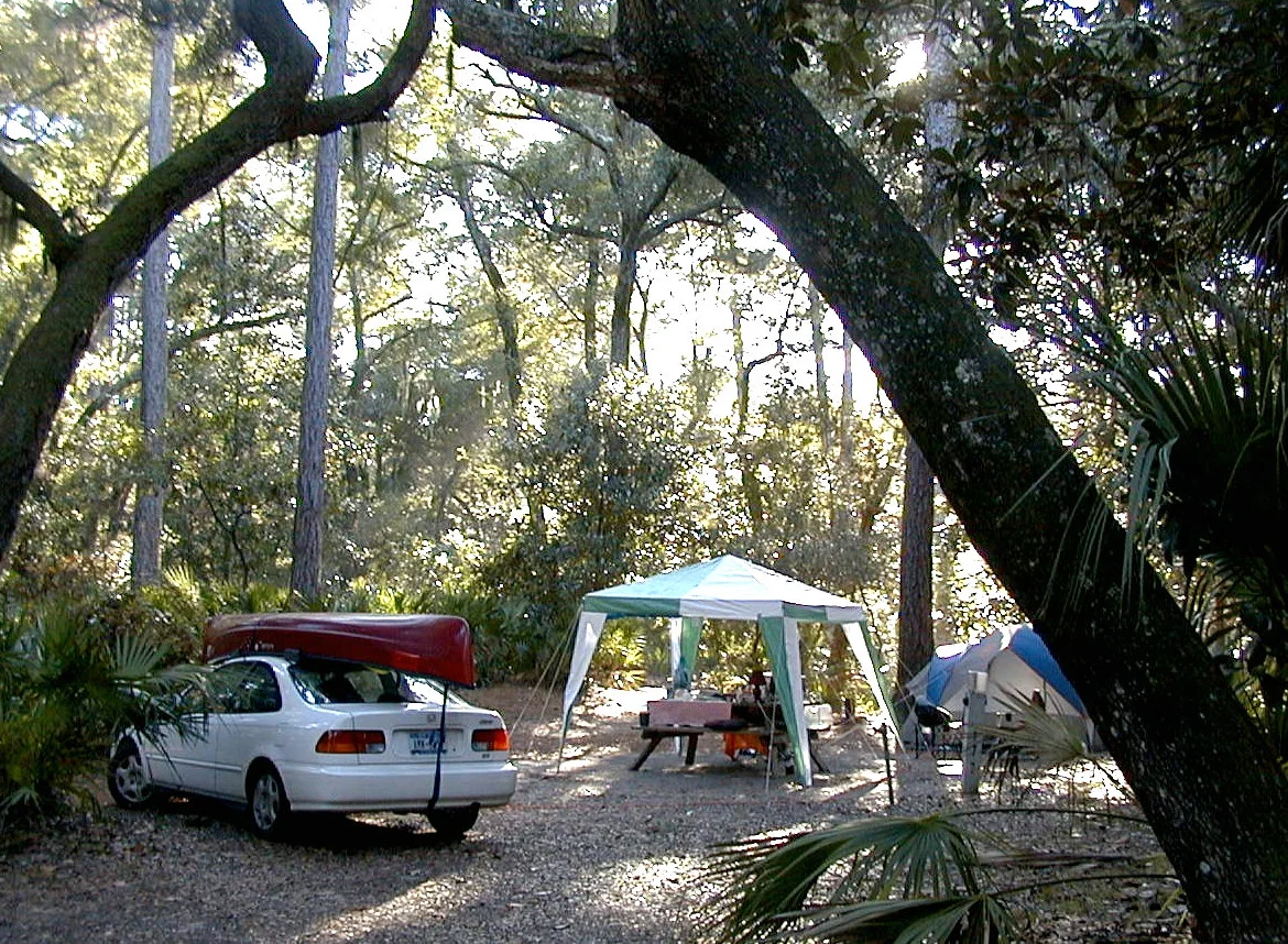 A car parked at a wooded campsite with a canopy and tent, the kind of trip a DIY sleeping platform is built for