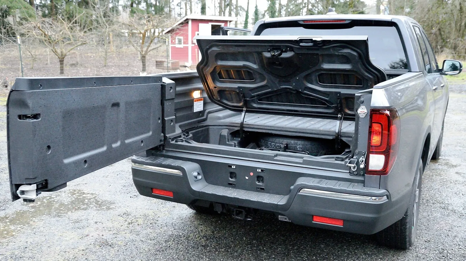 A grey second-generation Honda Ridgeline with its dual-action tailgate swung open to the side, revealing the lockable in-bed trunk and spare tyre behind the composite pickup bed used for camping storage.