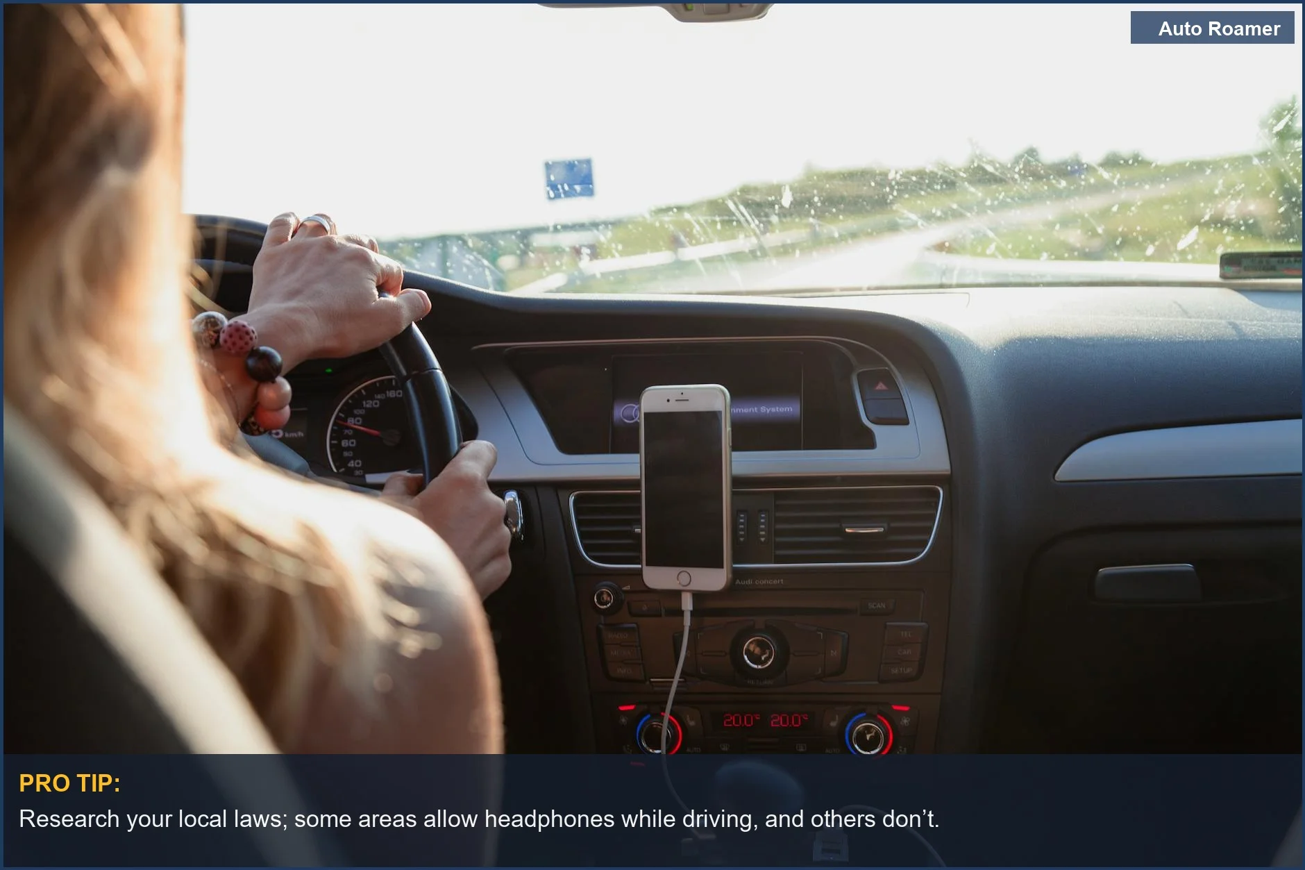 Woman driving a car on a sunny day, with a phone on the dashboard, reflecting on headphone use legality.