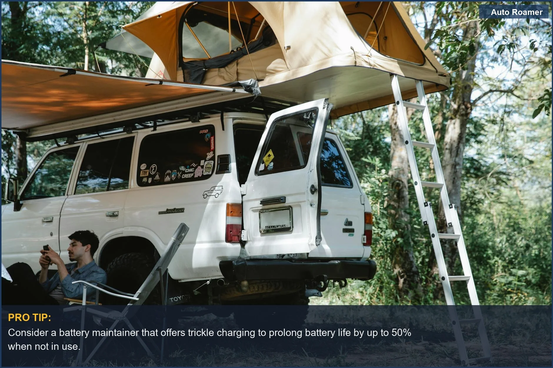 Man relaxing with smartphone near off-road vehicle and rooftop tent in a forest setting.