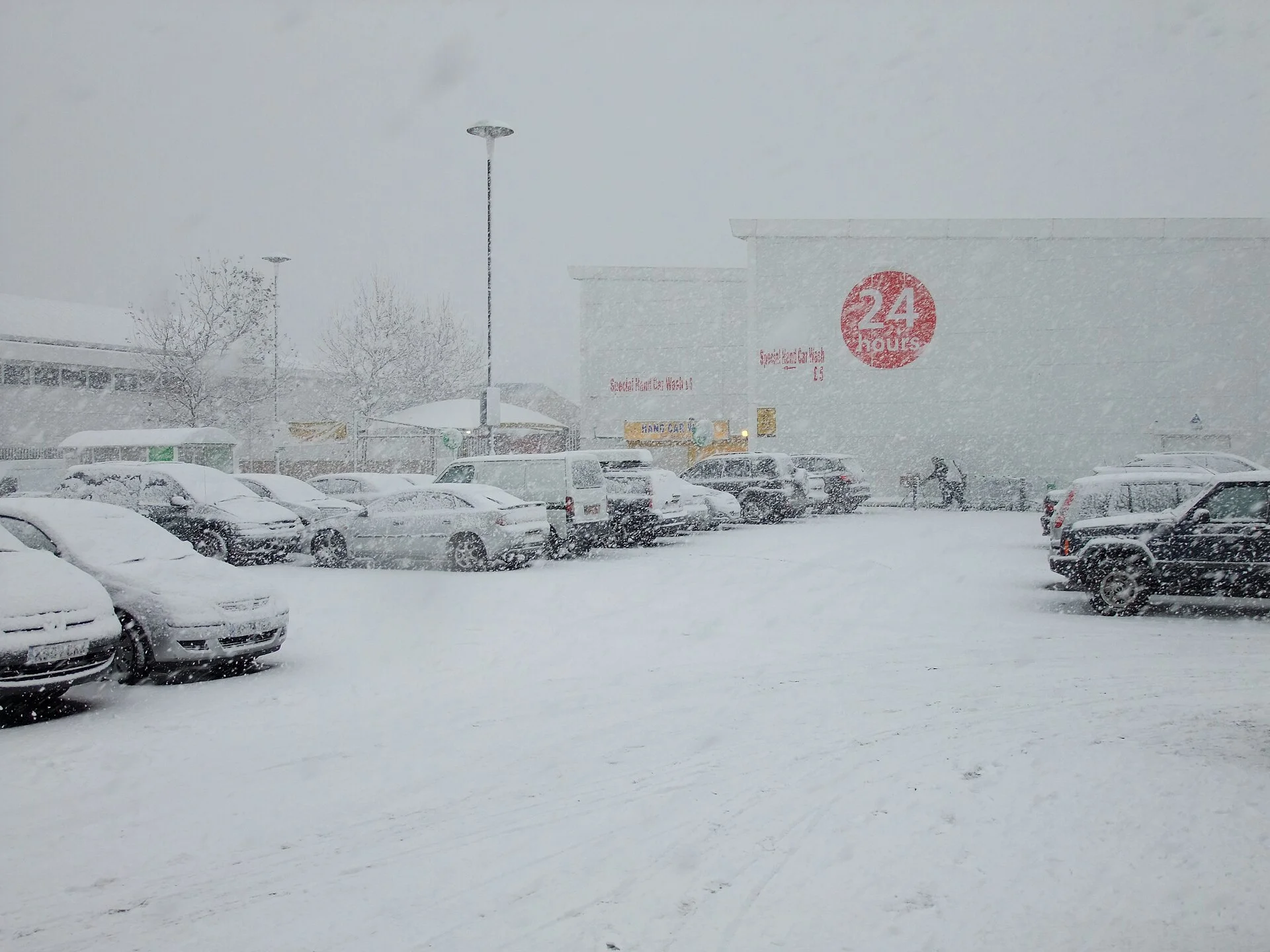 A supermarket car park during heavy snowfall, its parked cars buried under fresh snow — the kind of freezing overnight conditions this guide warns can make sleeping in a car dangerous.