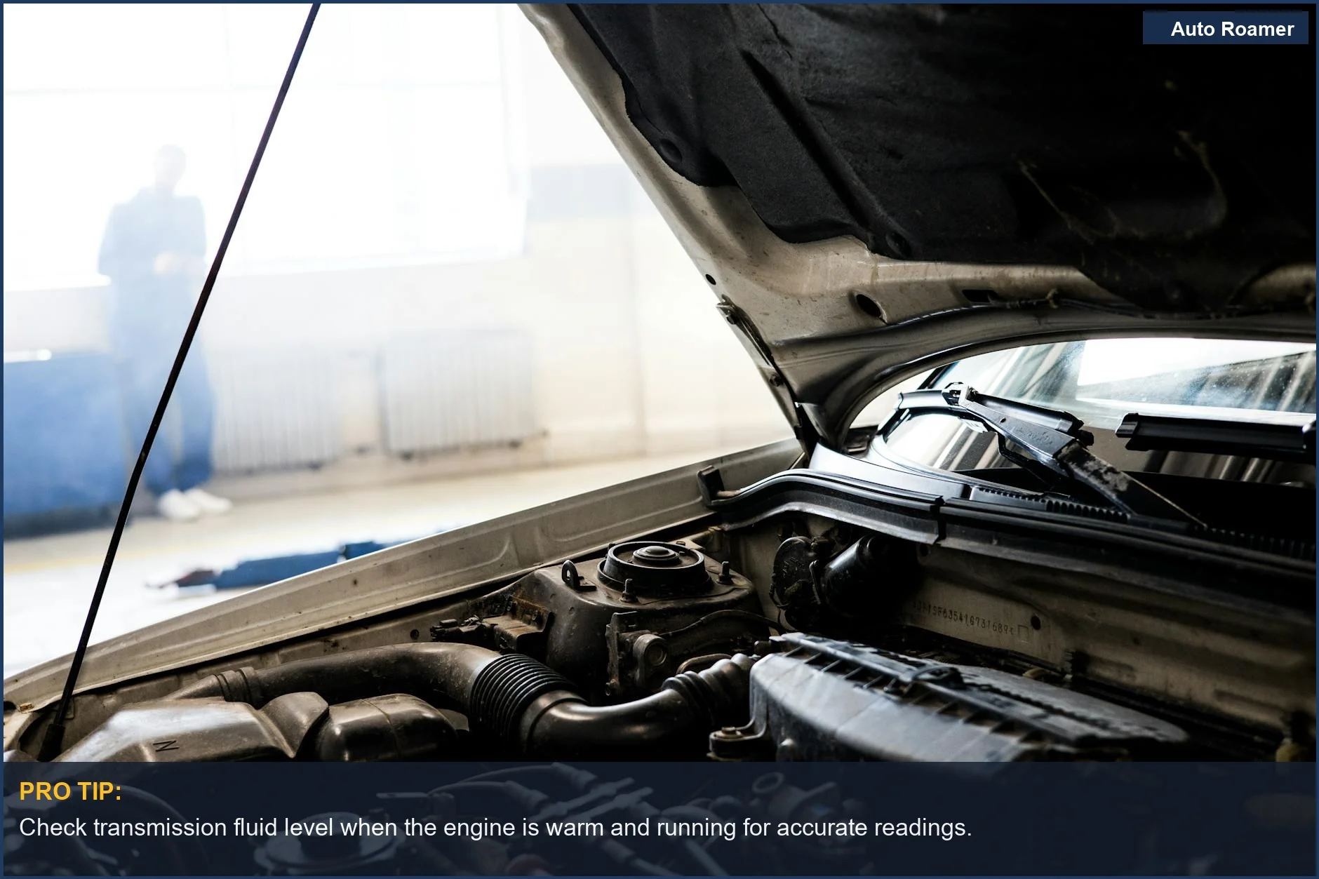 Mechanic checking transmission fluid level under a car's open hood in a well-lit garage.