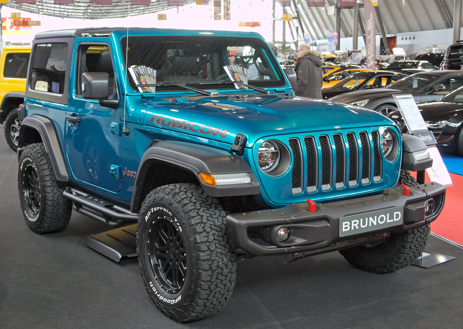 Teal two-door Jeep Wrangler Rubicon, current JL generation, black hardtop, front three-quarter view on an indoor auto-show stand, with all-terrain tyres.