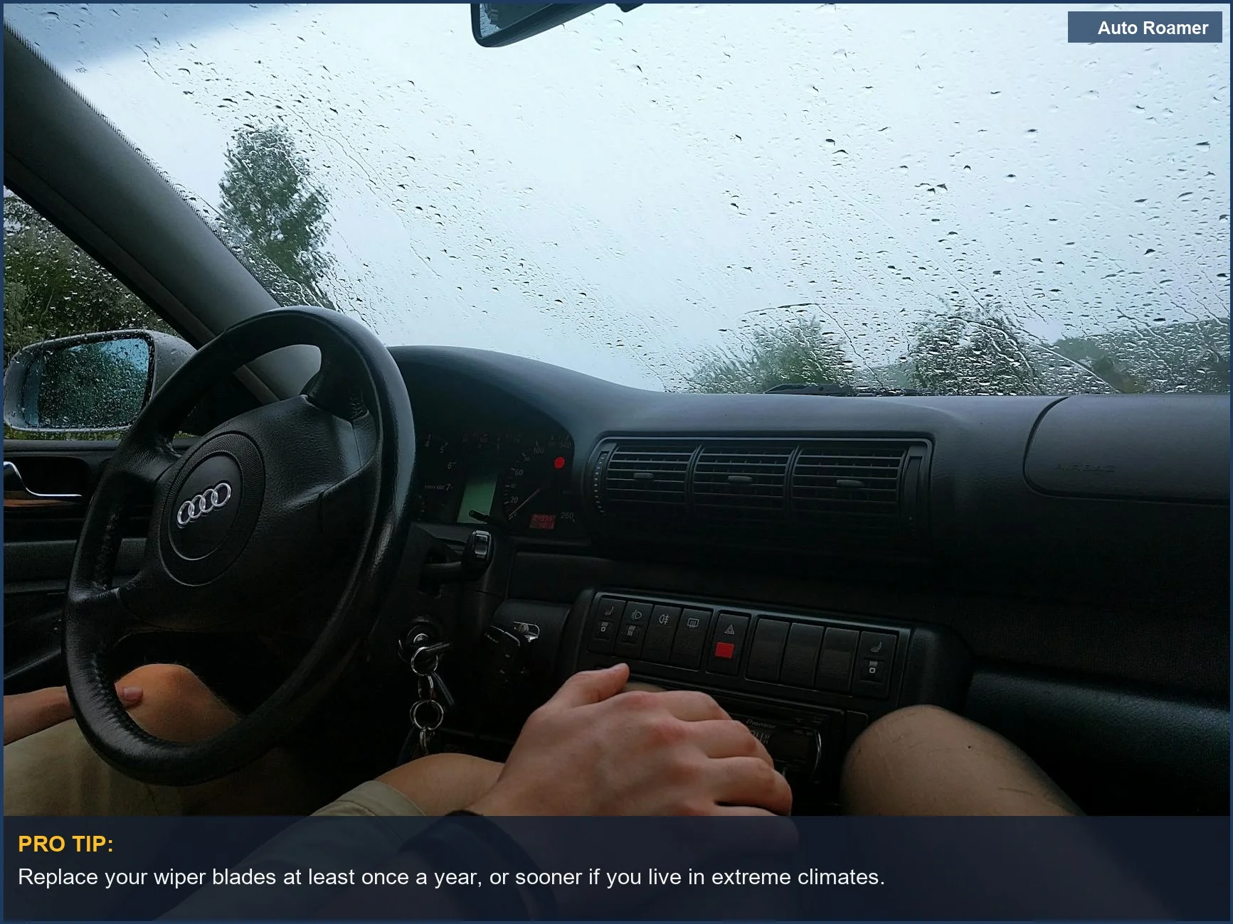 Steering wheel and dashboard in a car on a rainy day, with raindrops on the windshield, signaling need for wiper change.